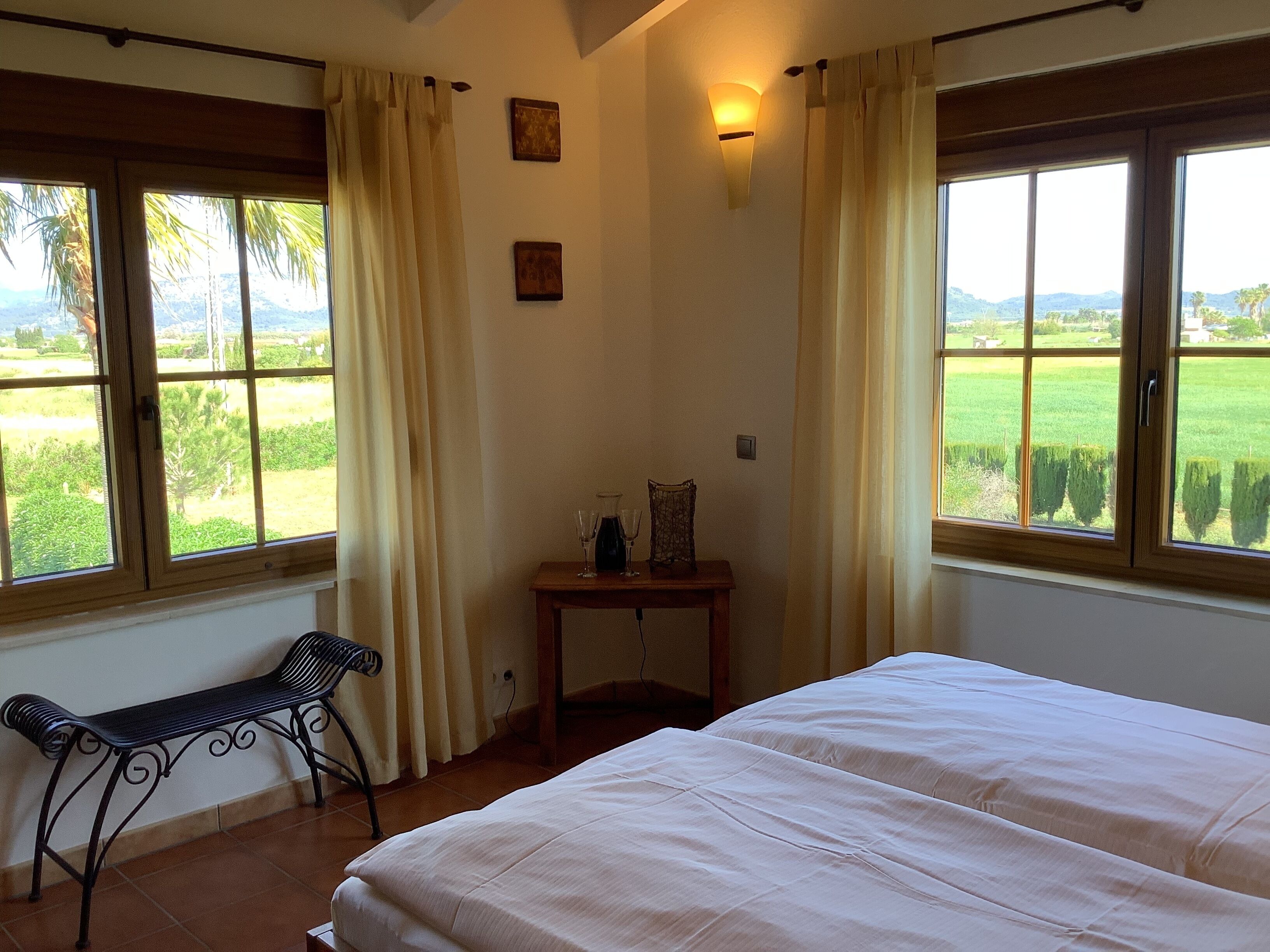 Un dormitorio con cama, un banco, una mesa con vasos y cortinas.