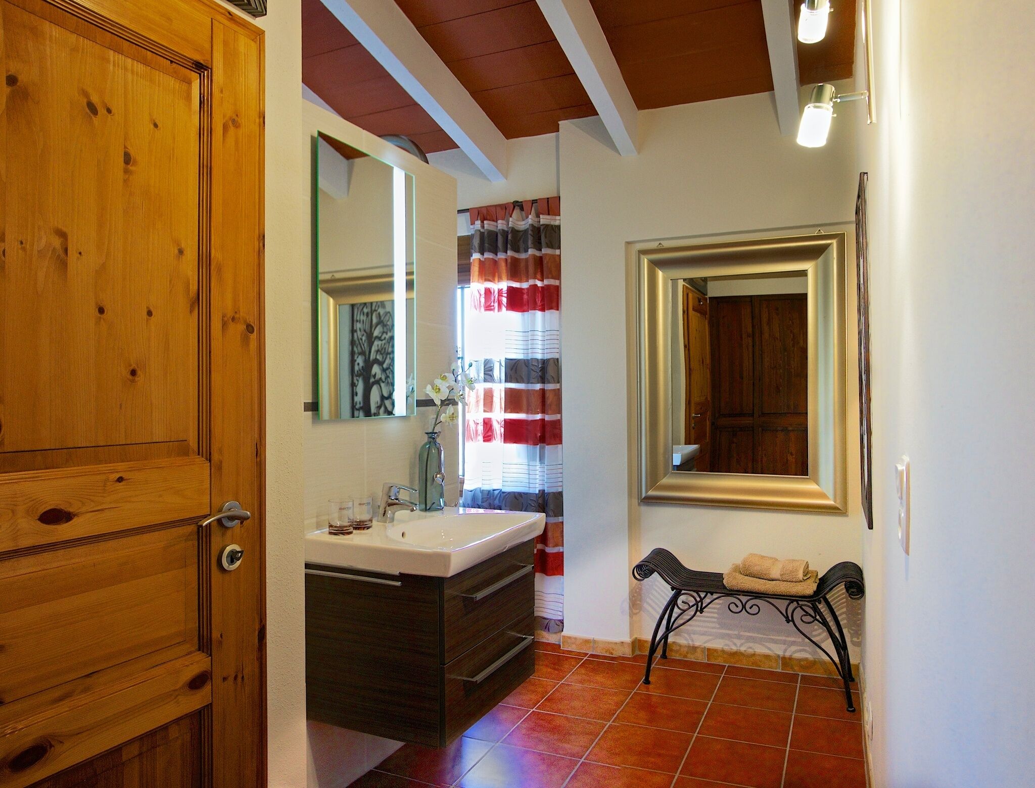 Une salle de bain avec une porte en bois, un lavabo avec un miroir, un banc avec des serviettes et une fenêtre avec des rideaux.