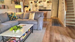 A cozy living room with a sectional sofa, a coffee table with books and plants, a dining area with a wooden table and chairs, and a kitchen in the background. - Water View 1 mi from Oak Bluffs 10 min Walk to Beach (Oak Bluffs)