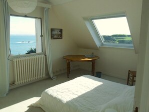 A bedroom with a bed, a table, a chair, a radiator, and a window with a view of the sea.