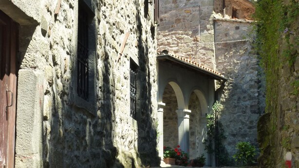 A narrow cobblestone street flanked by stone buildings with arched doorways and a small garden area with potted plants.