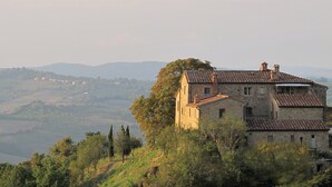 Ein Gebäude aus Stein mit Ziegeldach, umgeben von Bäumen und Hügeln im Hintergrund.