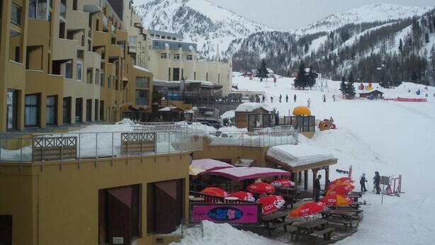 Un hotel con varios pisos y balcones, un complejo de esquí al fondo y un paisaje montañoso nevado.