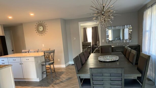 Een ruime eethoek met een lange tafel en moderne stoelen, een keuken met witte kasten, en een decoratieve wandklok.