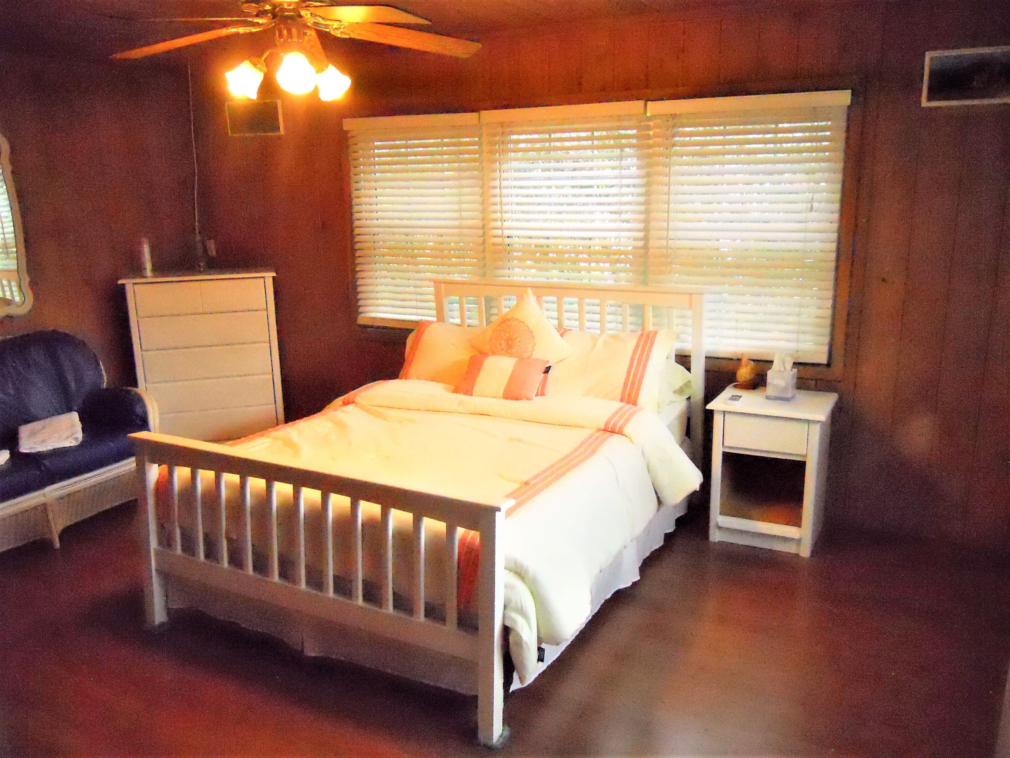 Een slaapkamer met een bed, nachtkastje, dressoir, stoel en plafondventilator.