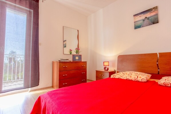 A bedroom with a large bed, a wooden dresser, a nightstand with a lamp, a mirror, and a window with curtains.
