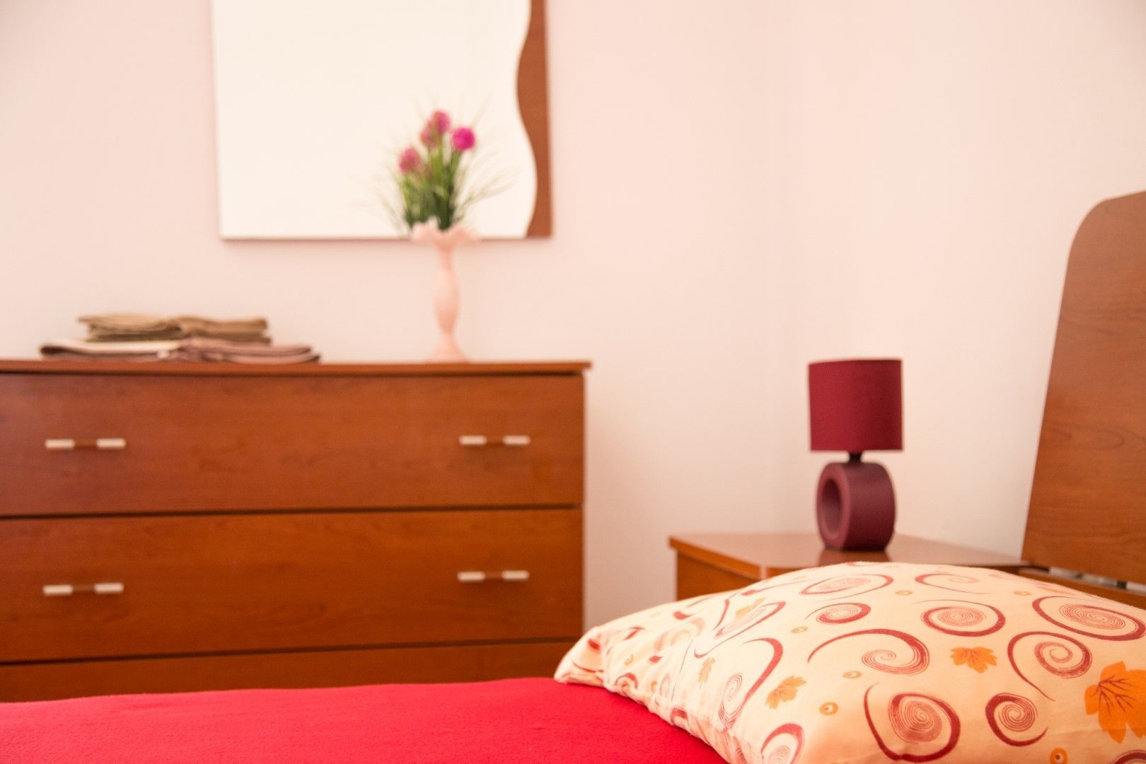 A bedroom with a wooden dresser, a bedside table with a lamp, and a bed with a red blanket and patterned pillow.