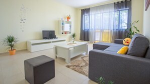 Un salon moderne avec un canapé gris, une table basse blanche et un téléviseur à écran plat posé sur un meuble blanc. Une grande fenêtre est agrémentée de rideaux légers, une plante en pot et une petite table sur laquelle sont disposés des objets décoratifs.