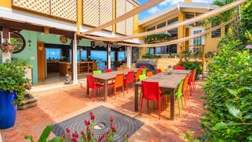 Un espace extérieur abrité, aménagé pour les repas, avec une table en bois et des chaises colorées, entouré de plantes en pot et offrant une vue sur la mer.