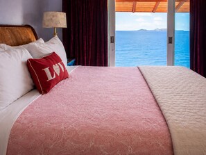 A bed with a patterned bedspread, a red pillow with 'LOVE' on it, and a view of the sea through a large window. - Cliffside Retreat | Infinity Pool, Yoga Deck, 9 BR (St. Thomas)