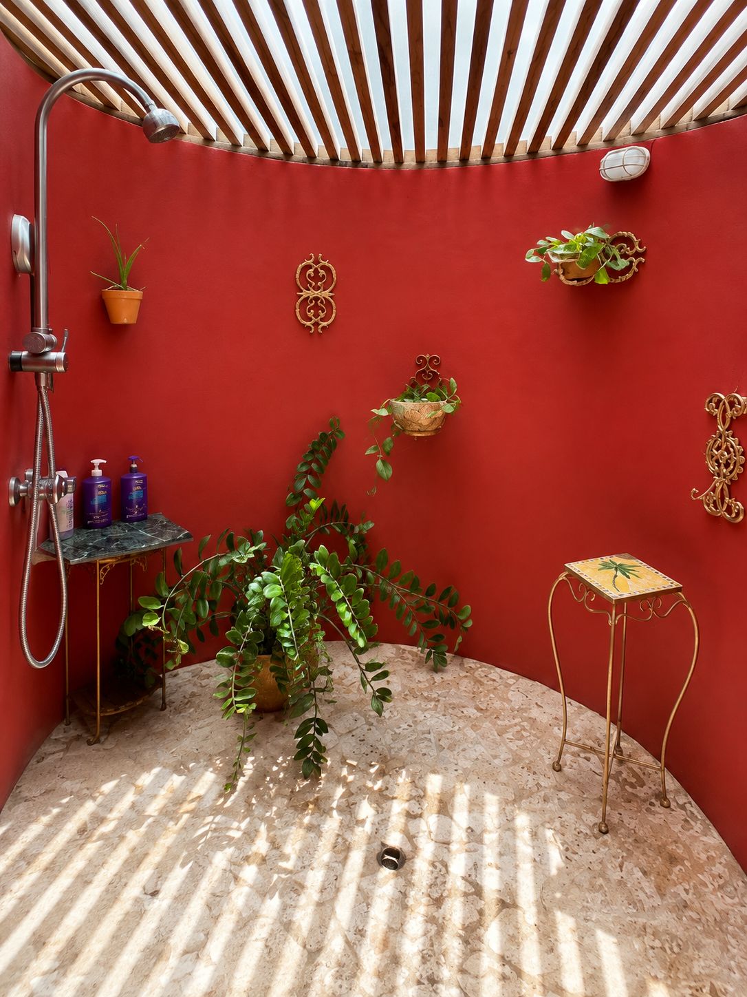 Une salle de bain aux murs rouges, équipée d’une douche, d’une petite table où sont posés les articles de toilette et agrémentée de plantes décoratives.