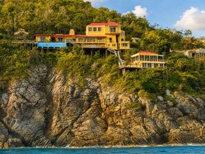 A multi-story building with a red roof, situated on a rocky cliff overlooking the sea. - Cliffside Retreat | Infinity Pool, Yoga Deck, 9 BR (St. Thomas)