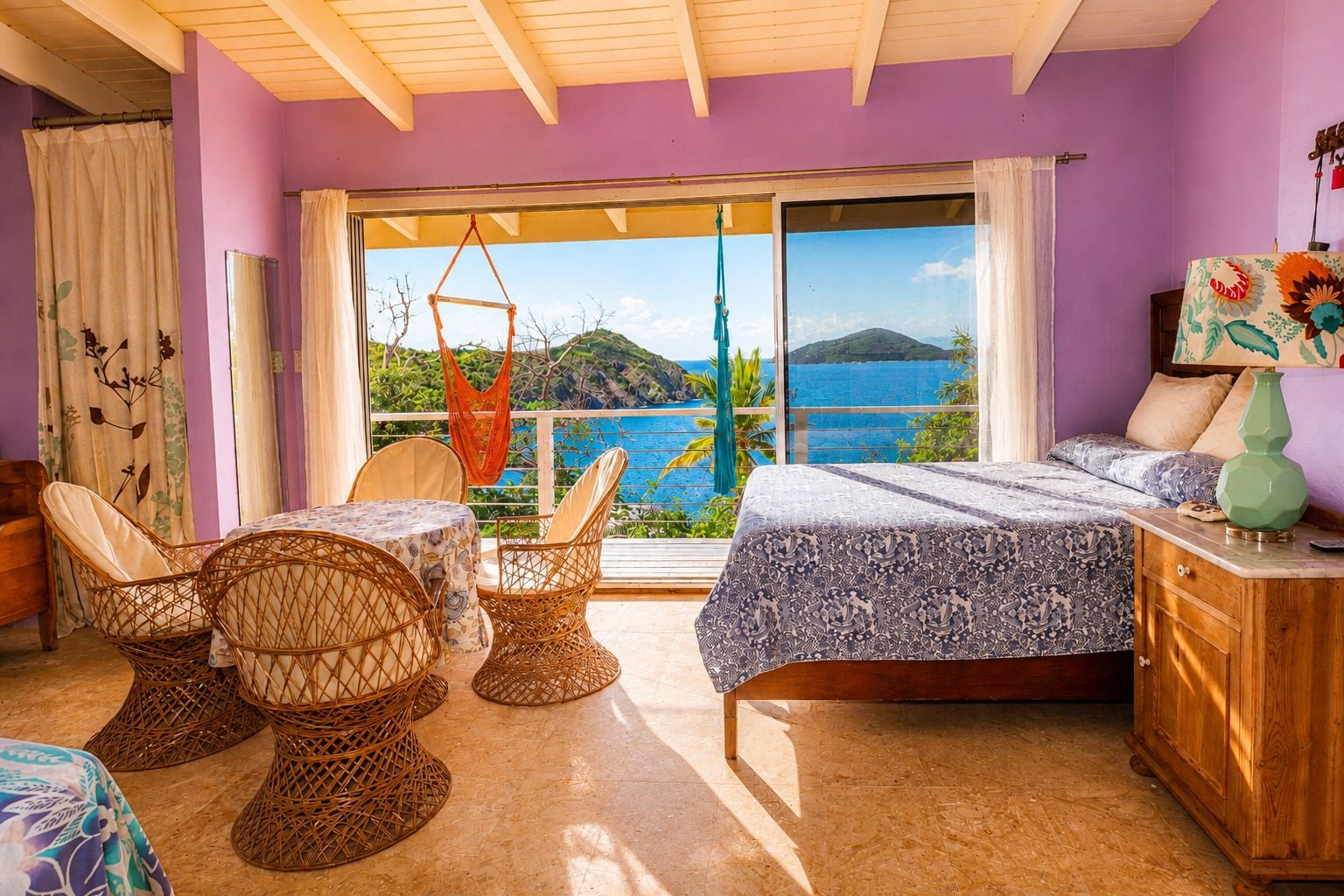 Ein Zimmer mit einem Bett, einem Nachttisch aus Holz, Korbstühlen, einem Tisch und Blick auf das Meer durch ein großes Fenster.