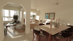 A spacious living area with a dining table, wicker chairs, a television, and a balcony with large windows. - Beautifully decorated apartment! Maulf Hill - Ocean View! 150m2 (Guarujá)