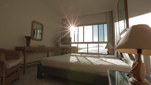 A bedroom with a bed, a bedside table, a lamp, a dresser, a mirror, and a window with a view of the outdoors. - Beautifully decorated apartment! Maulf Hill - Ocean View! 150m2 (Guarujá)