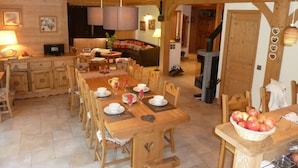 Un comedor de madera acogedor con una mesa larga dispuesta para cuatro personas, rodeada de sillas. Se observa una chimenea al fondo y una cesta de manzanas sobre la mesa.