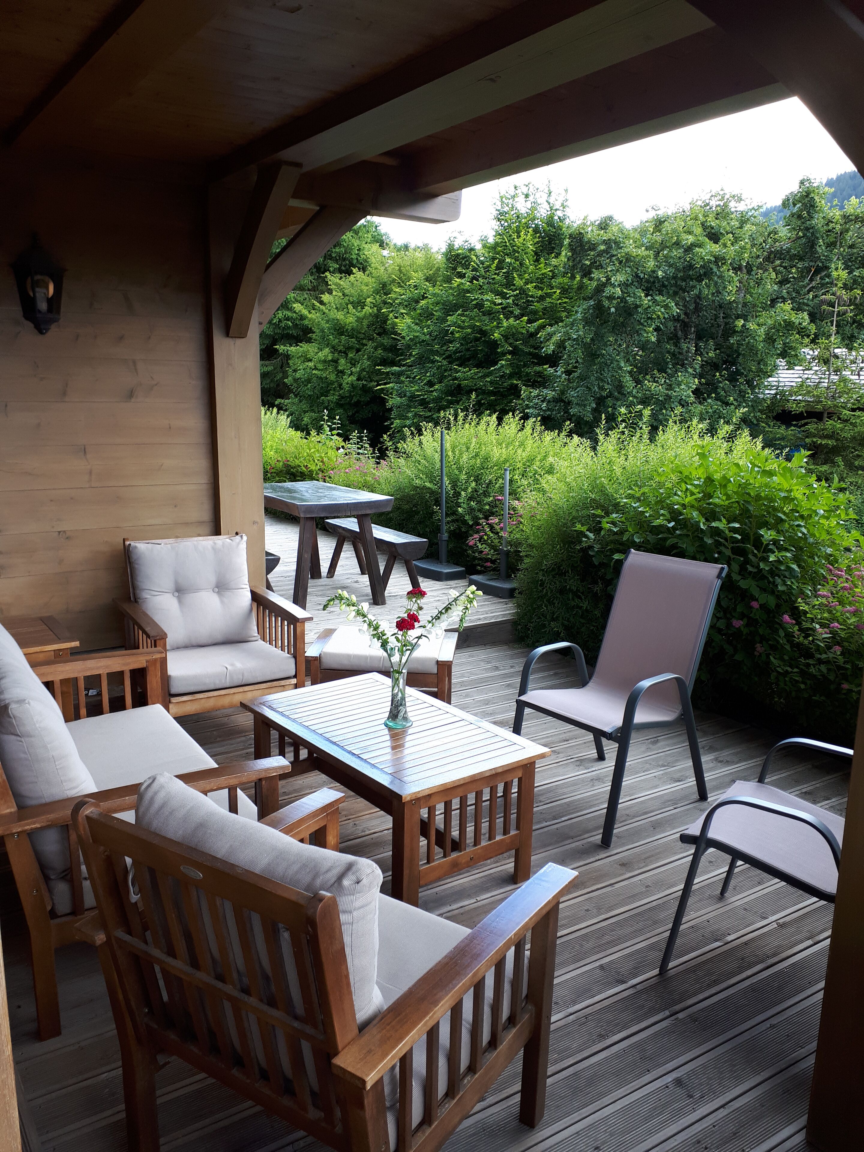 Une terrasse en bois, agréée de meubles d’extérieur, notamment des chaises et une table, et entourée d’une végétation luxuriante.