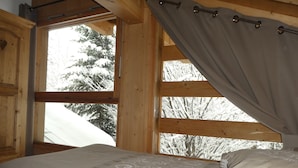 Un dormitorio de cabaña de madera con una cama, una ventana con cortinas y vistas a árboles cubiertos de nieve.