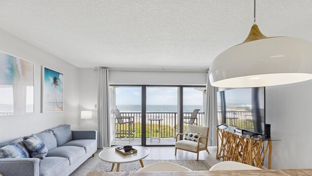 Ein modernes Wohnzimmer mit einer Couch, einem Couchtisch und Sesseln, einem Esstisch mit Tischdekoration sowie einem Balkon mit Meerblick.
