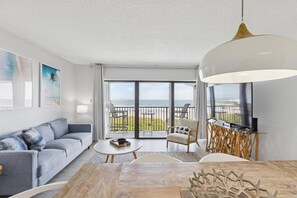 A modern living room with a sofa, coffee table, and chairs, a dining table with a centerpiece, and a balcony with a view of the sea.