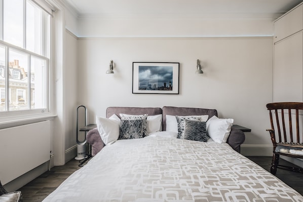 A neatly arranged bedroom with a large bed, a framed cityscape picture, and a chair.