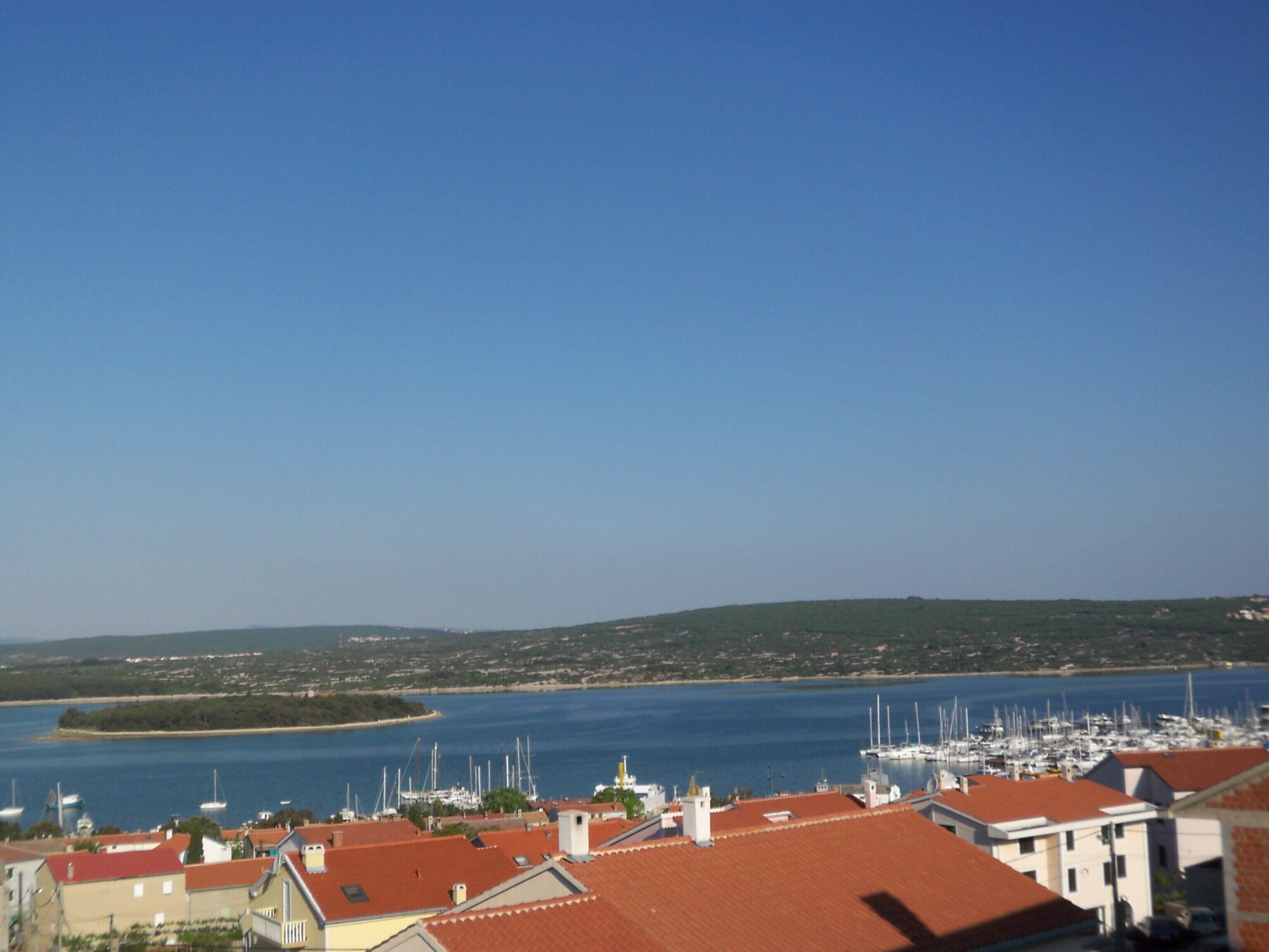 Uma vila costeira com edifícios de telhados vermelhos, um porto com barcos e um céu azul límpido.