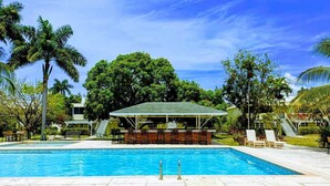 Una piscina con zona de bar techada, rodeada de palmeras y un cielo despejado.