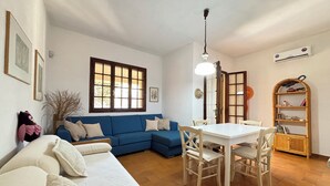 A living room with a blue sofa, white couch, and a dining table with chairs. There is a window with wooden frames, a ceiling fan, and an air conditioning unit on the wall. - Private villa with garden for two families !!100m from Solanas beach! 🏖 (Solanas)