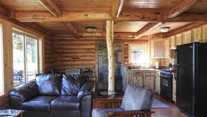 A cozy cabin interior with a wooden ceiling, a fireplace, a kitchen area, and leather seating. - Cabins At Hay Creek Gorgeous Log Cabin Stunning Views of Glacier National Park (Polebridge)