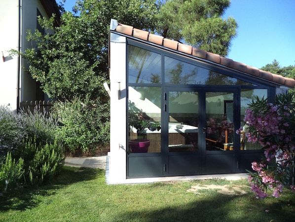 A modern glass conservatory with a tiled roof, surrounded by lush greenery and flowering plants.
