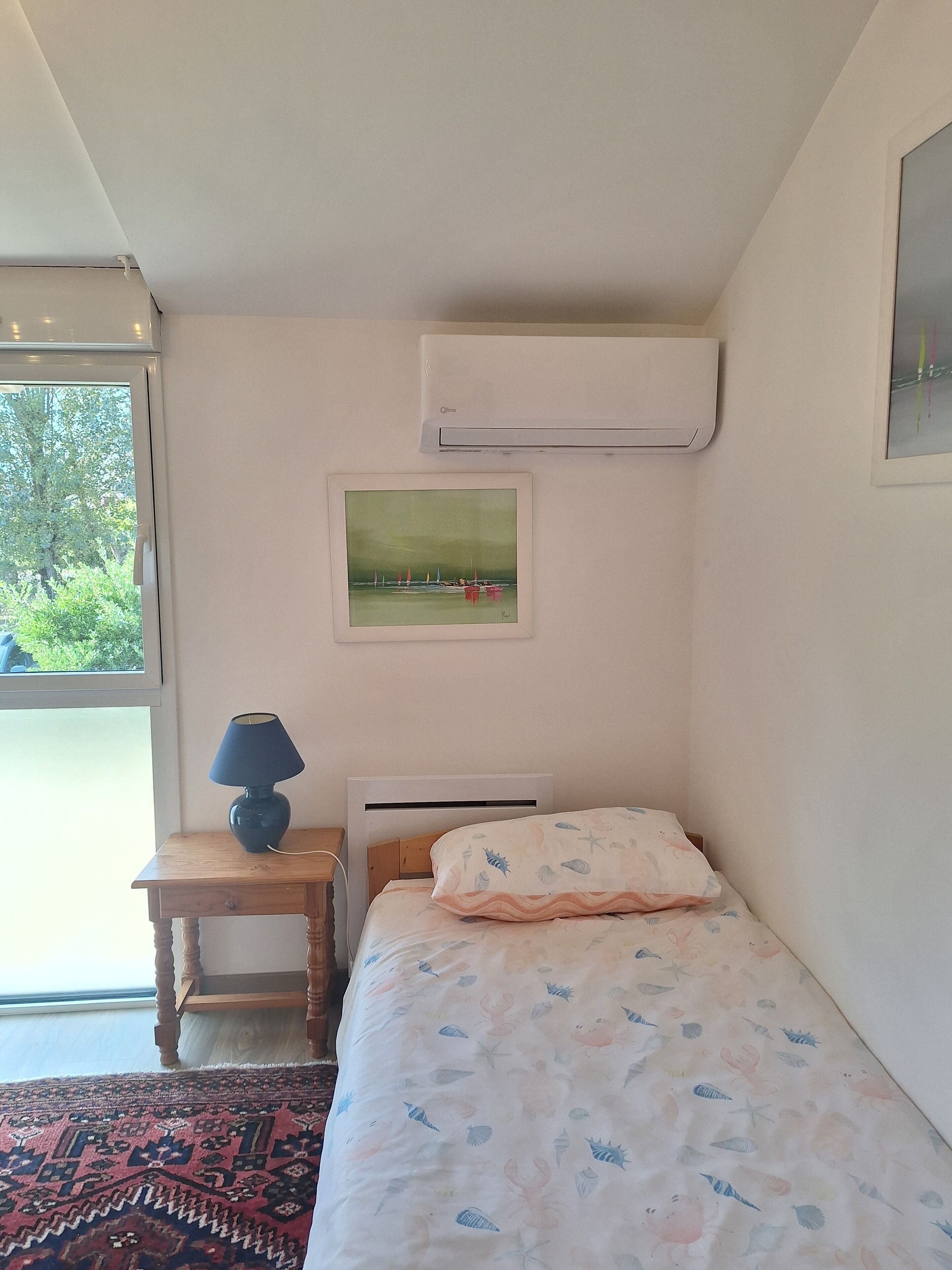 Une chambre à coucher avec un lit, une table de chevet, une lampe, un climatiseur, un tableau encadré et une vue par la fenêtre.