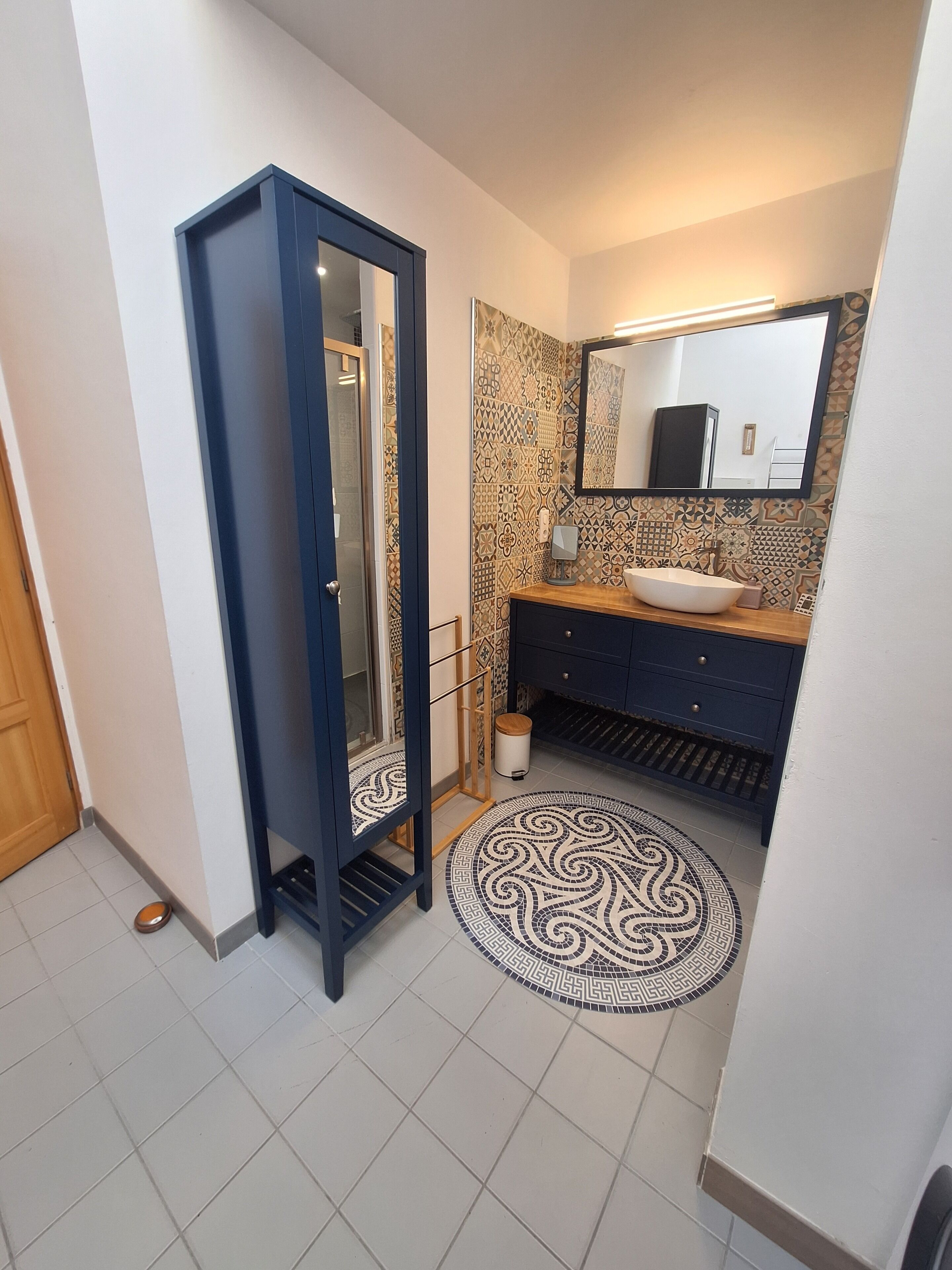 Ein Badezimmer mit gefliestem Boden, einem runden Teppich mit Muster, einem Waschbecken mit Spiegel und einem Kleiderschrank.
