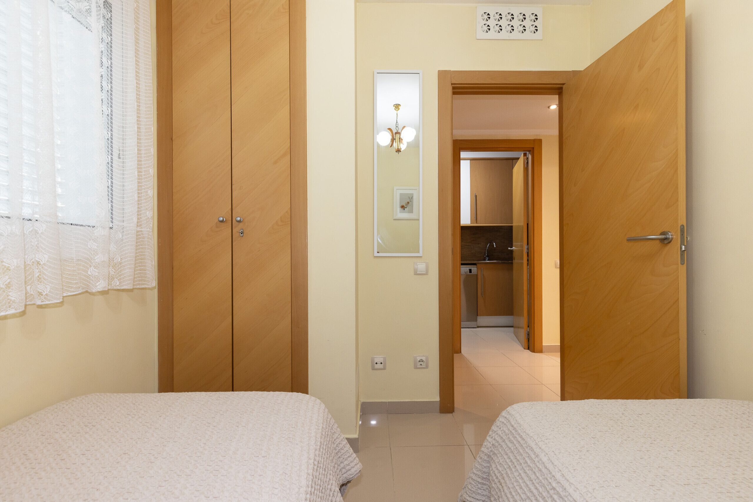 Quarto com duas camas, armários de madeira, um espelho e um corredor que dá para uma cozinha.