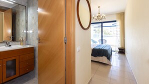 A hotel room with a wooden door, a large mirror, a bed with a blue bedspread, a chandelier, and a view of the outside.