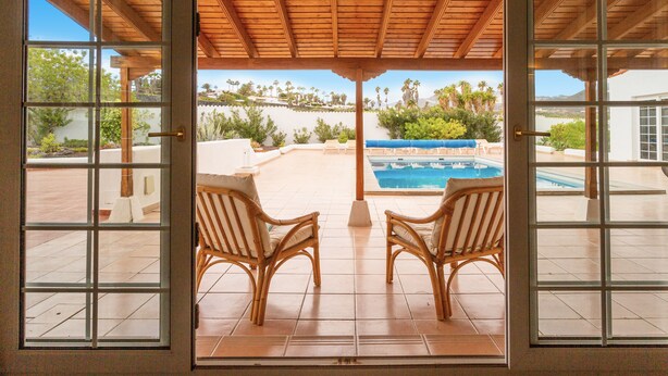 A covered patio with two wicker chairs facing a swimming pool and a view of a landscaped area with trees and a building in the background.