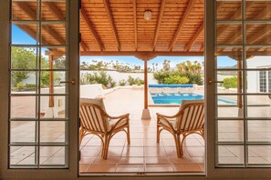 Un patio techado con dos sillones de mimbre mirando a una piscina y vistas de una zona ajardinada con árboles y un edificio al fondo.