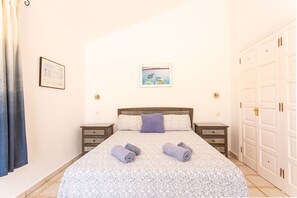Una cama bien hecha con una colcha estampada con motivos florales, dos toallas enrolladas, una única almohada morada y un cuadro en la pared.