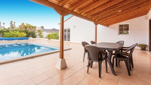 A covered outdoor area with a pool, a table with chairs, and a view of the surrounding landscape.
