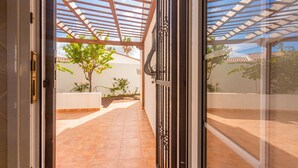 A covered outdoor area with a tiled floor, a pergola, and a view of a tree and building in the background.