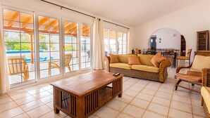 A spacious living area with a large sliding glass door leading to a poolside area, a wooden coffee table, a wicker sofa, and a dining set.