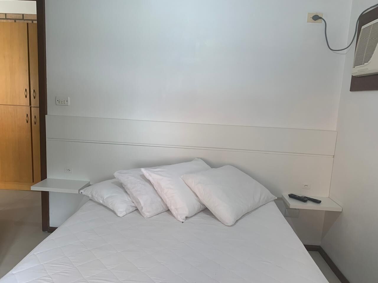Una cama bien hecha con ropa blanca y almohadas, una mesita de noche con control remoto y un equipo de aire acondicionado instalado en la pared.