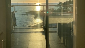 Une vue depuis un balcon avec une porte vitrée, donnant sur un coucher de soleil sur l’eau, avec des bateaux et une côte lointaine.