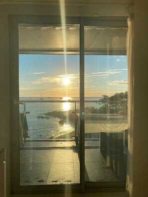 Uitzicht vanaf een balkon met een glazen deur, op een zonsondergang boven het water met boten en een verre kustlijn.