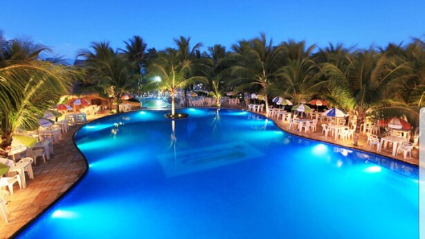 A large, curved swimming pool surrounded by palm trees and lounge chairs, with a clear blue sky in the background. - Apartment in Fortaleza, CE, Praia do Futuro, 3 bedrooms, 100m2 with 3 air conditioners, Wi-Fi. (Fortaleza)