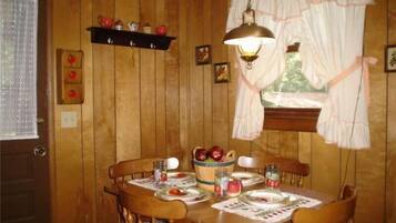 Une salle à manger en bois avec une table dressée pour quatre personnes, agrémentée d’une nappe à motifs floraux, d’assiettes et d’un pichet. Une étagère au-dessus de la table contient des pommes rouges et un petit vase.
