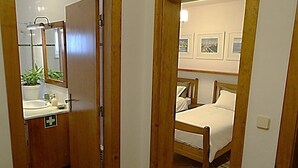 Un dormitorio con cama, mesita de noche y velador. Se ve una ventana con vista a un paisaje urbano.