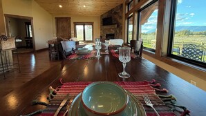 A dining area with a wooden table set for four, a stone fireplace, and large windows offering a view of a green landscape. - Cozy Cabin | 20 MI to YNP | Walk to Henry’s Lake (Island Park)