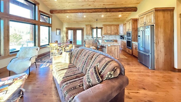 A spacious living area with a large sofa, wooden ceiling beams, and a kitchen with wooden cabinets and appliances. - Cozy Cabin | 20 MI to YNP | Walk to Henry’s Lake (Island Park)