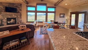 A spacious living area with a stone fireplace, large windows, a dining table, and a kitchen island. - Cozy Cabin | 20 MI to YNP | Walk to Henry’s Lake (Island Park)