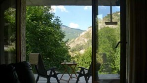 A room with a view of a mountainous landscape through a glass door. There is a small table with two chairs and a window. - Spacious apartment with large terrace in nature (Allos)